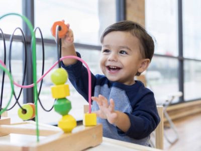 istockphoto-1056360056-612x612 An adorable toddler boy sits at a table in a doctor's waiting room and reaches up cheerfully to play with a toy bead maze.