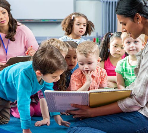 Female teacher giving a lesson to nursery students. They are sitting on the floor and there is a teacher taking notes.