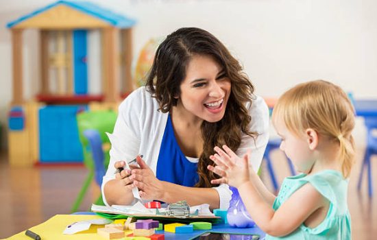 Cheerful female Hispanic teacher praises adorable blonde Caucasian little girl on accomplishing a project. The preschooler claps along with the teacher.